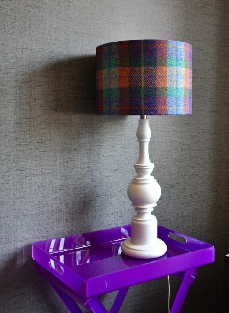 A purple, orange and blue check drum shade made from Harris Tweed on a white wooden lamp base on a purple perspex table. The lampshade is availble to order through Telescope Style.