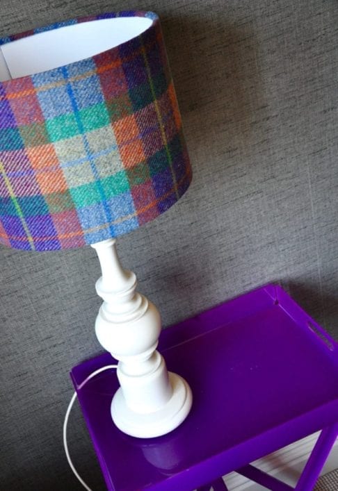 A brightly-hued check dru lampshade handmade from Harris tweed fabric sat on a purple perspex side table against a grey wallpapered wall. Available to order through Telescope Style.