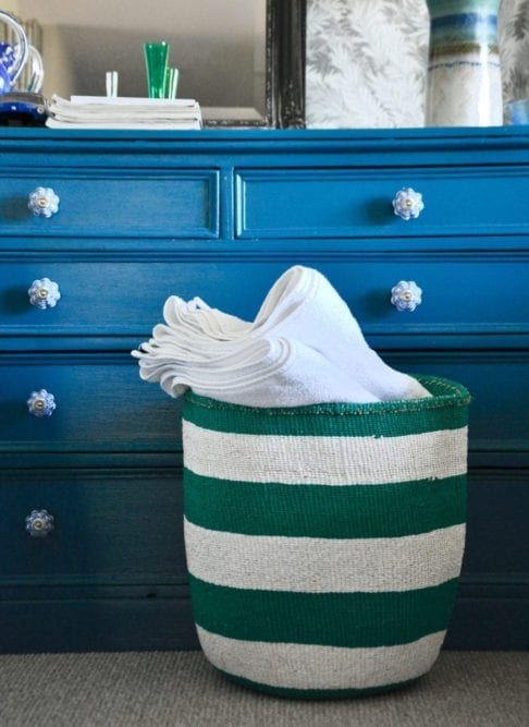 A jade green and white striped, hand-woven basket filled with towels in front of a teal chest of drawers.