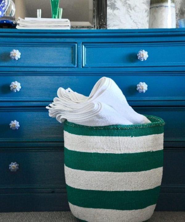 A jade green and white striped, hand-woven basket filled with towels in front of a teal chest of drawers.