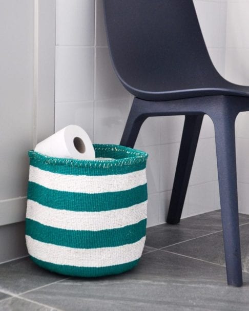 A jade green striped basket used as a loo roll holder with a chair in the background.