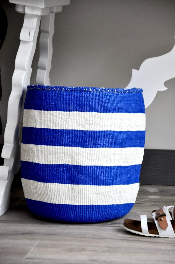 A blue and white striped, hand-woven basket, handmade in Kenya and displayed here as storage under a white refectory table.