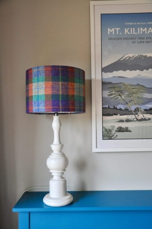 A Harris tweed lampshade teamed with a white wooden base on a teal, painted sideboard with a retro travel print on the wall behind.