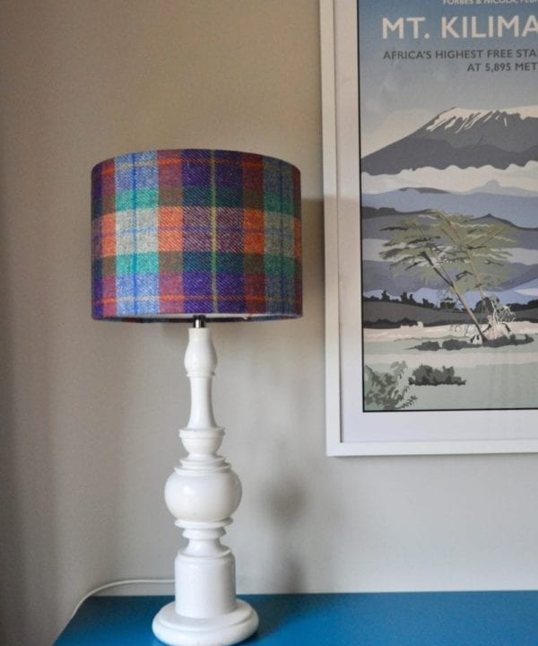 A Harris tweed lampshade teamed with a white wooden base on a teal, painted sideboard with a retro travel print on the wall behind.