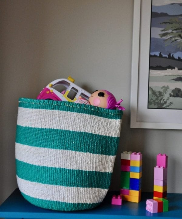 A stripy jade woven basket with colourful kids toys and lego inside on a teal sideboard.
