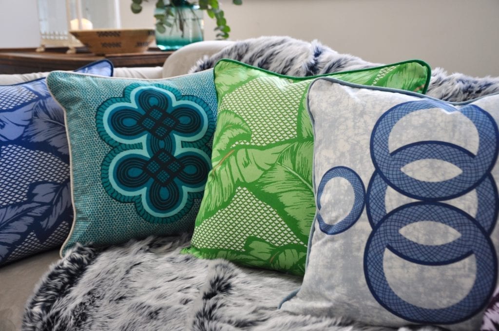 A vibrant selection of aqua, blue and green African-inspired cushions arranged on a grey, faux fur throw on a sofa.