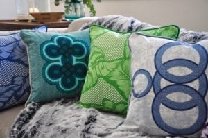 A vibrant selection of aqua, blue and green African-inspired cushions arranged on a grey, faux fur throw on a sofa.