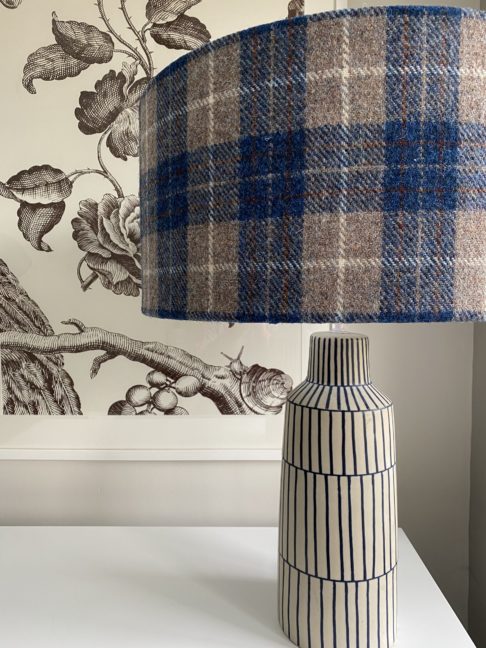 A blue check drum shade handmade from Harris Tweed on a blue and white lamp base with a black floral print in the background.