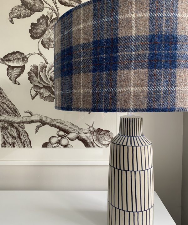 A blue check drum shade handmade from Harris Tweed on a blue and white lamp base with a black floral print in the background.