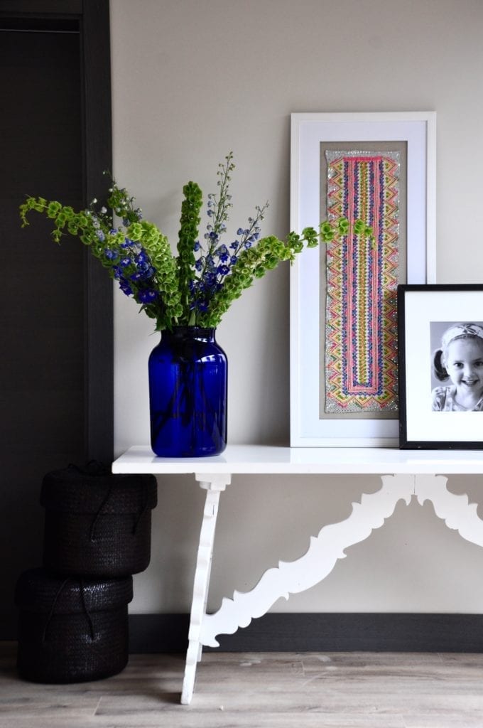 A neon 'colour pop' Thai framed textile displayed on a white painted side table with a large blue glass vase of Bells of Ireland and Delphiniums.