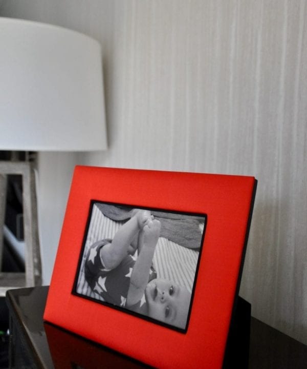 A Thai-silk, red, photograph frame with a picture of a baby next to a floor lamp and against a pale, strié-wallpapered backdrop.