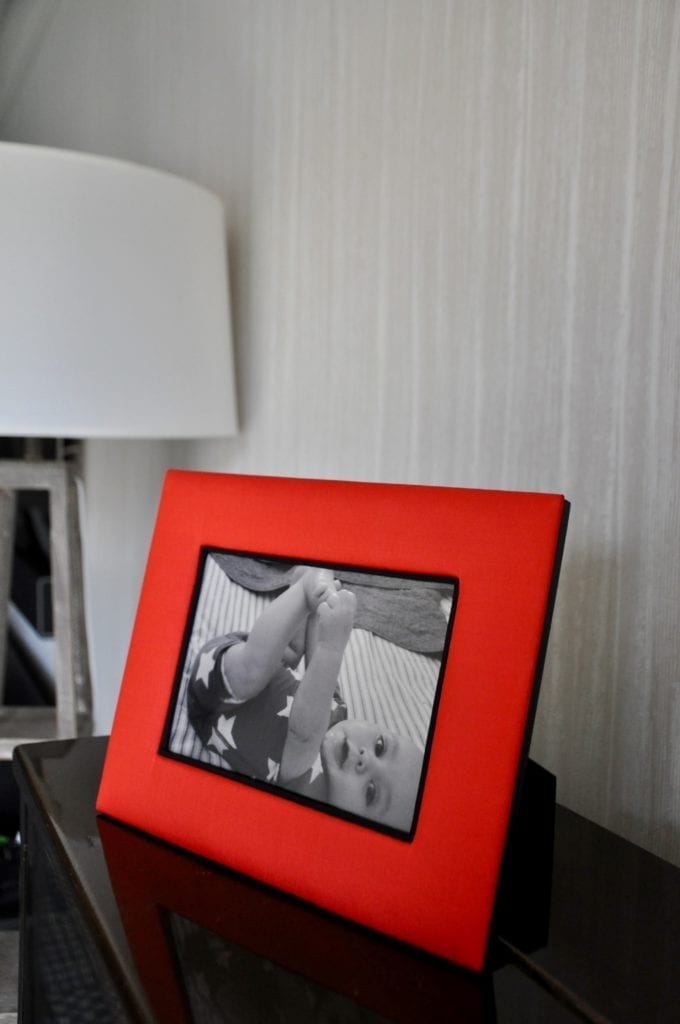 A Thai-silk, red, photograph frame with a picture of a baby next to a floor lamp and against a pale, strié-wallpapered backdrop.