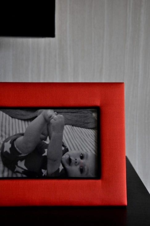 A Thai-silk, red, photograph frame with a picture of a baby against a pale, strié-wallpapered backdrop.