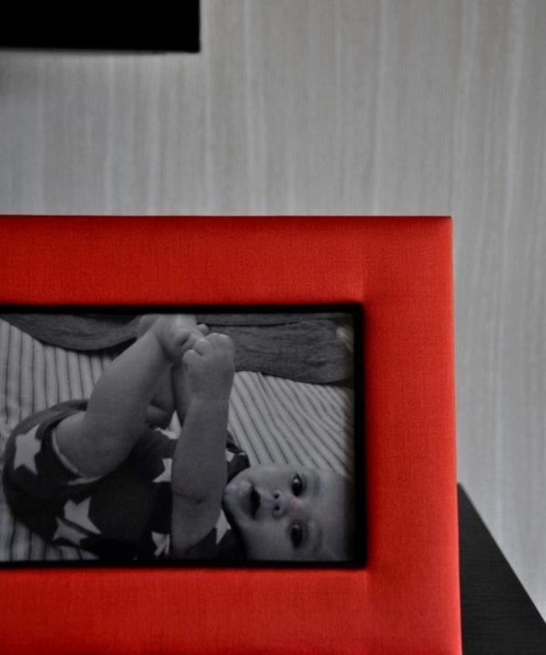 A Thai-silk, red, photograph frame with a picture of a baby against a pale, strié-wallpapered backdrop.