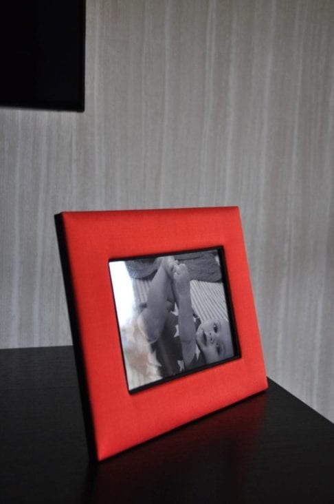 A Thai-silk, red, photograph frame with a picture of a baby against a pale, strié-wallpapered backdrop.