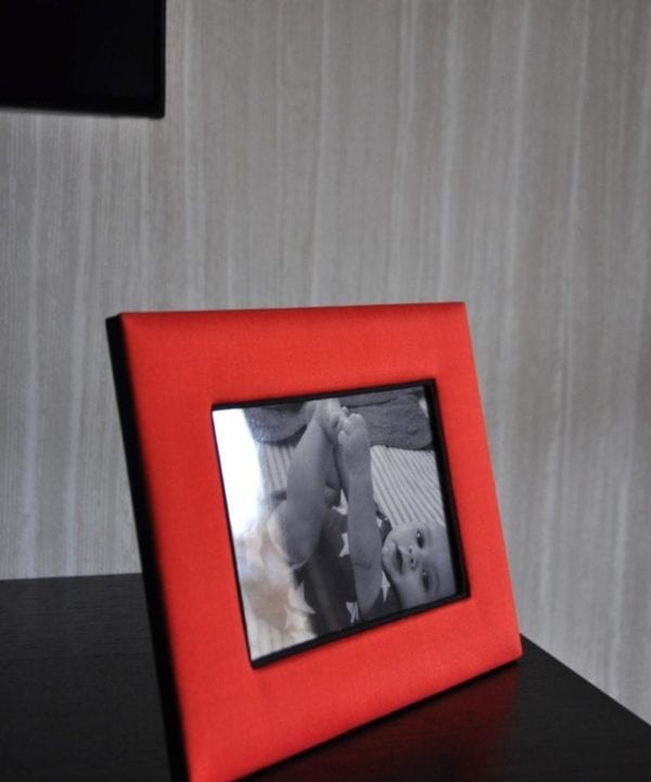 A Thai-silk, red, photograph frame with a picture of a baby against a pale, strié-wallpapered backdrop.