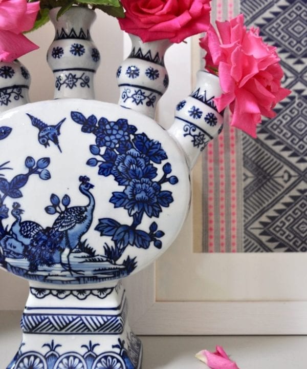 Detail of a Dutch-inspired blue and white painted tulip vase filled with pink roses on a ledge with a framed Thai textile behind.