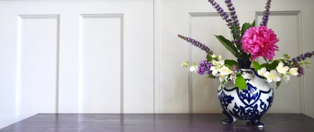 A landscape shot of a Dutch Delft tulip vase filled with summer blooms against a neutral, panelled backdrop.