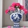 Front on shot of a Dutch-inspired blue and white painted tulip vase filled with pink roses on a ledge with a framed Thai textile behind.