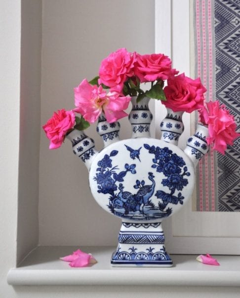 Front on shot of a Dutch-inspired blue and white painted tulip vase filled with pink roses on a ledge with a framed Thai textile behind.