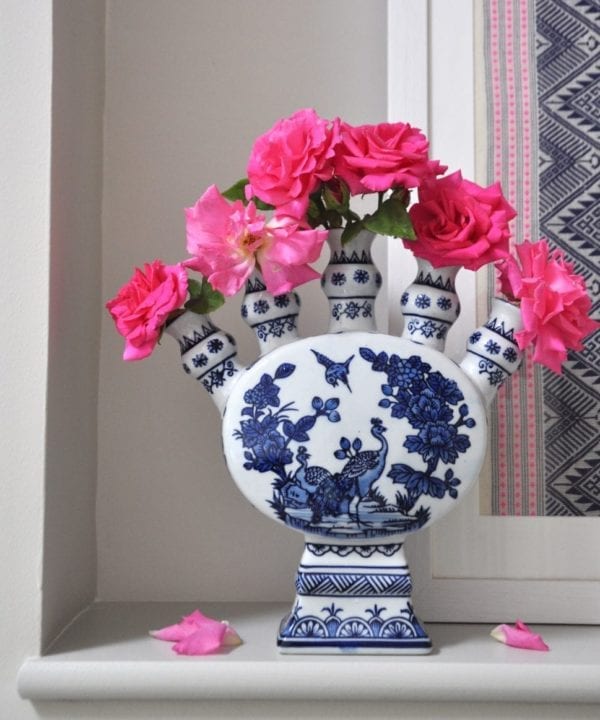 Front on shot of a Dutch-inspired blue and white painted tulip vase filled with pink roses on a ledge with a framed Thai textile behind.