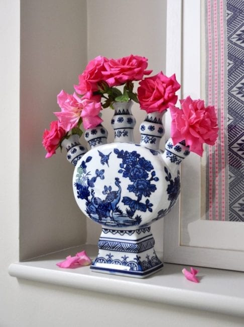 A Dutch-inspired blue and white painted tulip vase filled with pink roses on a ledge with a framed Thai textile behind.