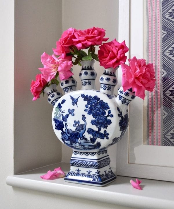 A Dutch-inspired blue and white painted tulip vase filled with pink roses on a ledge with a framed Thai textile behind.