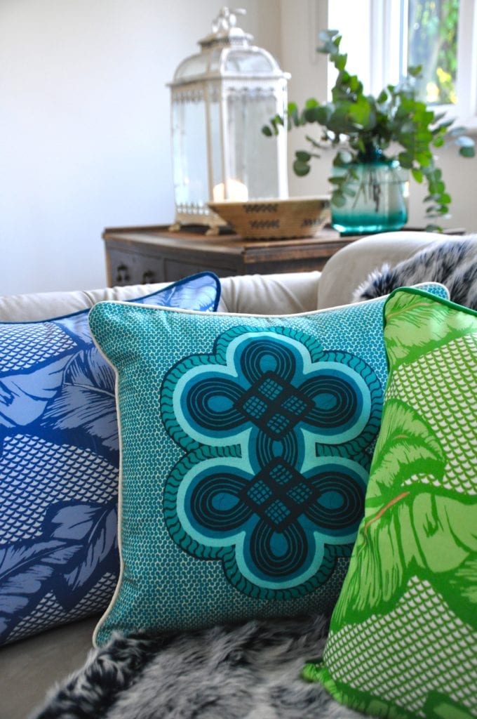 Bold, West African motifs on colourful cushions in blues and greens on a sofa with foliage and a candlelit lantern in the background.