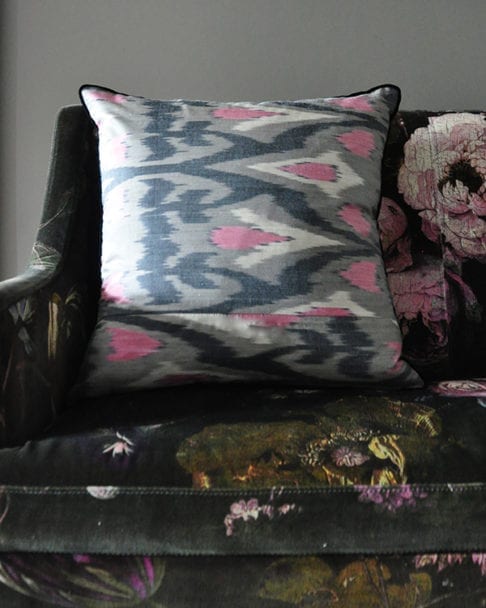 A horizontally-arranged, square, pink and warm, grey, silk ikat cushion with black accents sat on a floral velvet upholstered banquette.