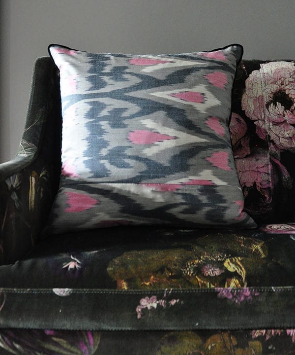 A horizontally-arranged, square, pink and warm, grey, silk ikat cushion with black accents sat on a floral velvet upholstered banquette.