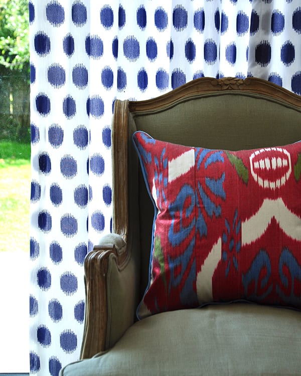 A colourful red, silk, ikat cushion on a French wing chair againstb a blue and white curtain with a view to the garden.