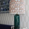 A statuesque patterned drum lampshade on a cream side table in front of geometric wallpaper.