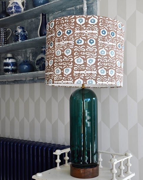 A statuesque patterned drum lampshade on a cream side table in front of geometric wallpaper.