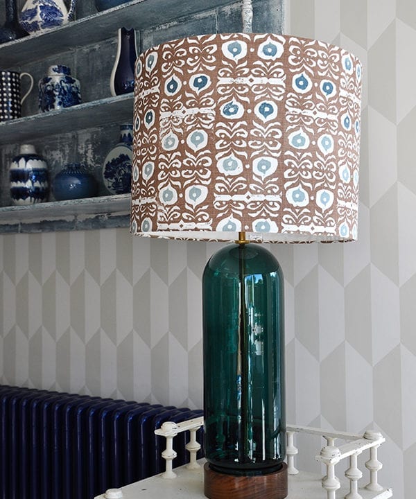 A statuesque patterned drum lampshade on a cream side table in front of geometric wallpaper.