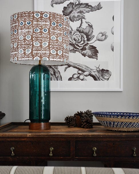 A teal blue and brown patterned lampshade on a wooden sideboard with a floral print in the background.