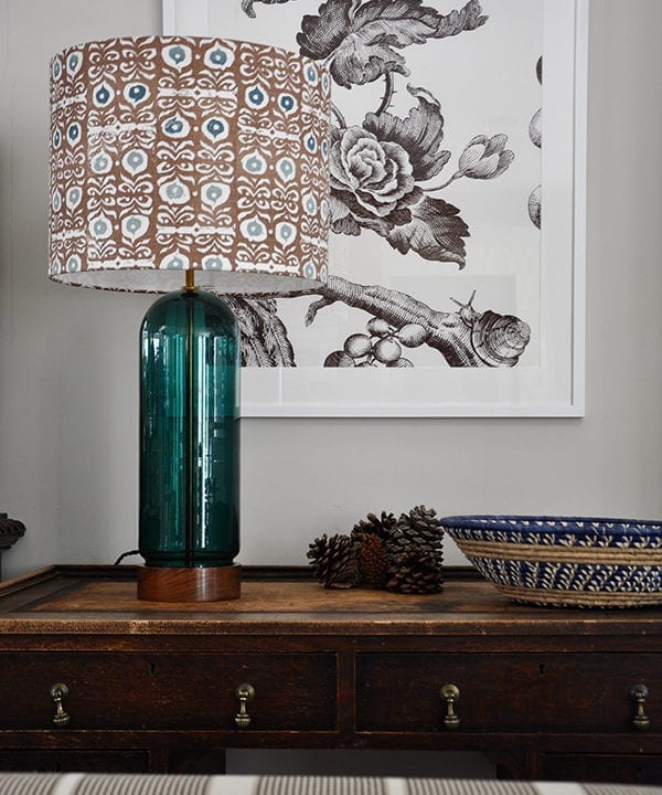 A teal blue and brown patterned lampshade on a wooden sideboard with a floral print in the background.