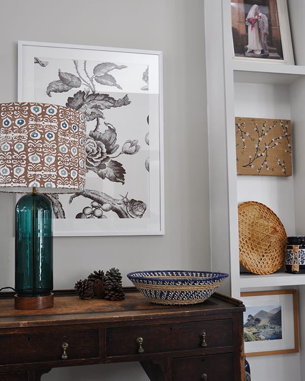 Shallow, built-in shelving next a wooden desk with a framed floral print above and a patterned lampshade on a teal glass base.