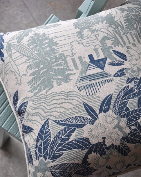 A pale blue floral cushion depicting an Arts & Crafts house in it's garden setting photographed on a pale turquoise chair.