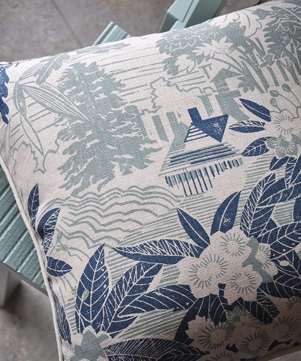 A pale blue floral cushion depicting an Arts & Crafts house in it's garden setting photographed on a pale turquoise chair.