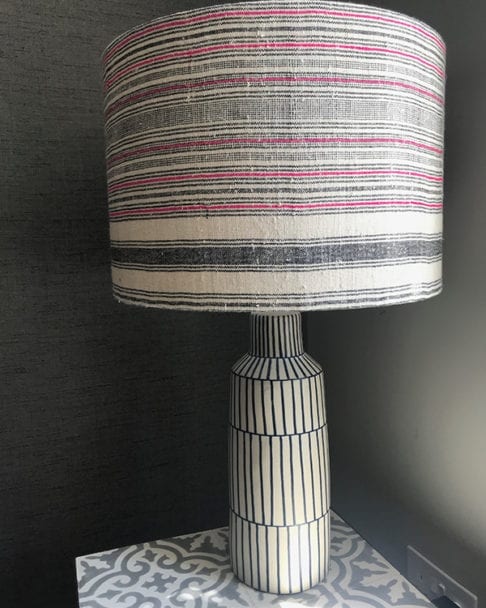 A striped pink, black and natural coloured vintage hemp lampshade handmade from a handwoven Thai textile on a Greek inspired ceramic lamp base on a grey patterned tiled side table.