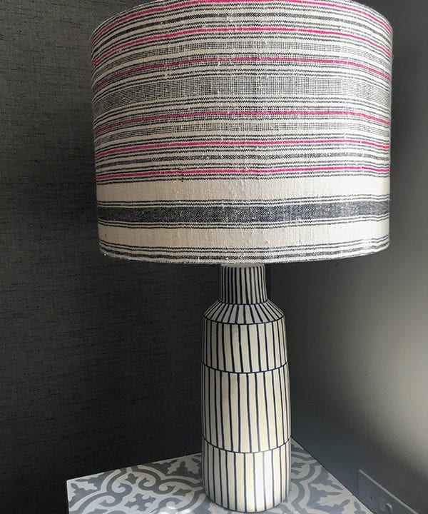 A striped pink, black and natural coloured vintage hemp lampshade handmade from a handwoven Thai textile on a Greek inspired ceramic lamp base on a grey patterned tiled side table.