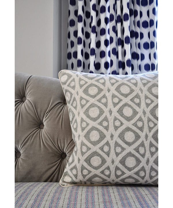 Cream and grey crewelwork cushions from Kashmir on a grey, buttoned, velvet sofa with a spotted blue curtain behind.