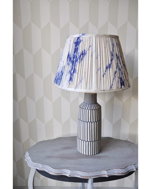 Indigo blue gathered silk lampshades in Japanese-style Shibori silk. The lampshade is sat on a painted, grey, side table with geometric wallpaper behind.