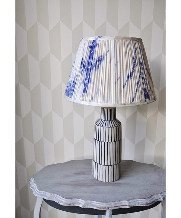 Indigo blue gathered silk lampshades in Japanese-style Shibori silk. The lampshade is sat on a painted, grey, side table with geometric wallpaper behind.