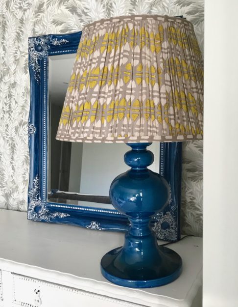 A lampshade in chartreuse and putty, block-printed in India on cotton voile, sits on a blue ceramic lamp base on a white sideboard.