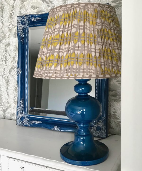 A lampshade in chartreuse and putty, block-printed in India on cotton voile, sits on a blue ceramic lamp base on a white sideboard.