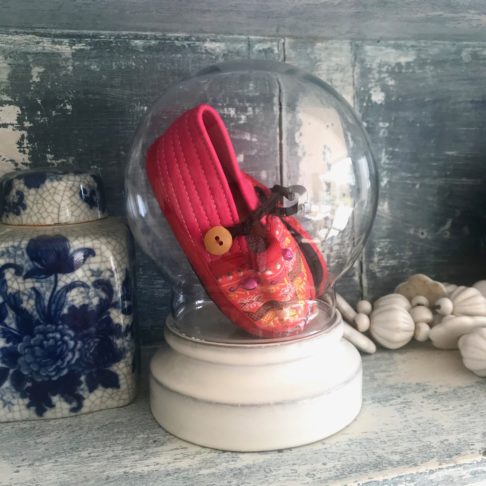 A round glass display dome holding pink, baby's Thai slippers showcased on pretty vintage shelving with blue ceramics.