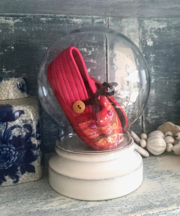 A round glass display dome holding pink, baby's Thai slippers showcased on pretty vintage shelving with blue ceramics.