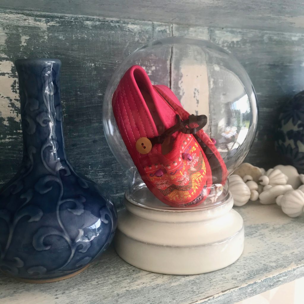 A bubble glass display dome holding pink, baby's Thai slippers showcased on pretty vintage shelving with blue ceramics.