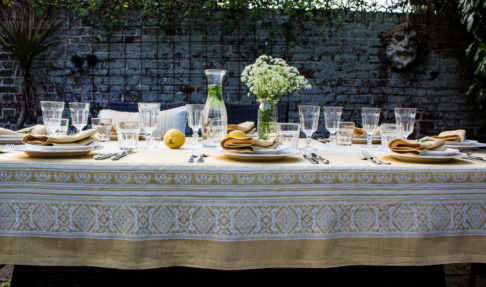 Yellow linen tablecloth with subtle motifs that reference Hungarian folk patterns. Draped over a table outside, set with glasses and tableware.